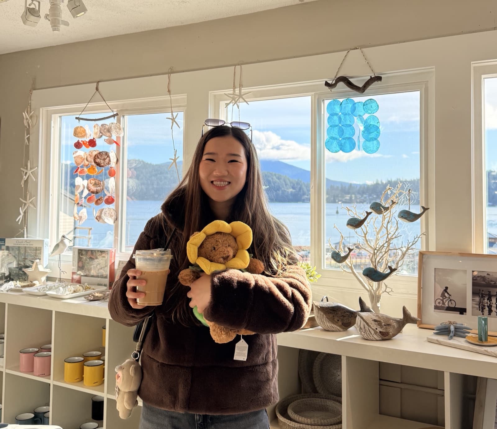 Katrina smiling in a seaside gift shop, holding an iced coffee and a sunflower teddy bear